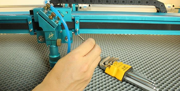 A technician wearing safety goggles and gloves checks the lenses and mirrors of a CO2 laser engraver, while a checklist is displayed on a nearby computer screen