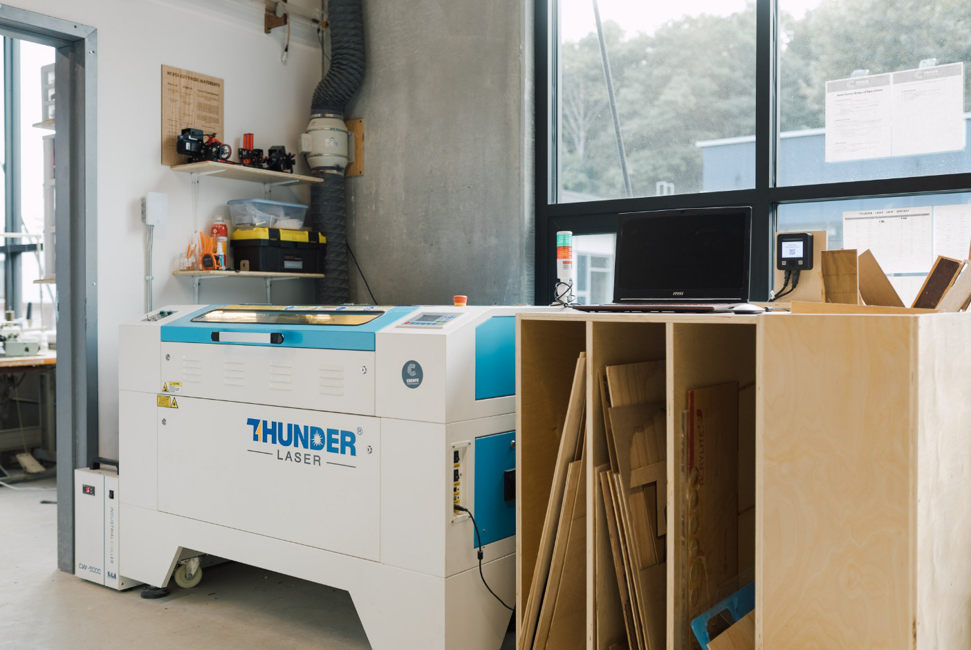 A CO2 laser engraver in a clean, well-lit workspace with safety equipment and a checklist posted nearby