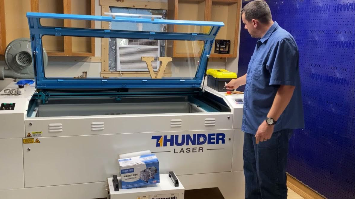 A person wearing safety goggles and gloves prepares vinyl sheet for laser cutting on a well-ventilated workbench with a laser cutter in the background