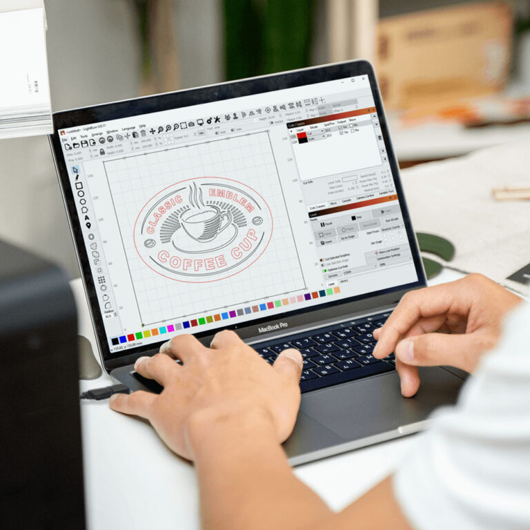 An artist sits at a computer, with a CO2 and fiber laser machine in the background. The artist is using Lightburn software to set up the machines for use